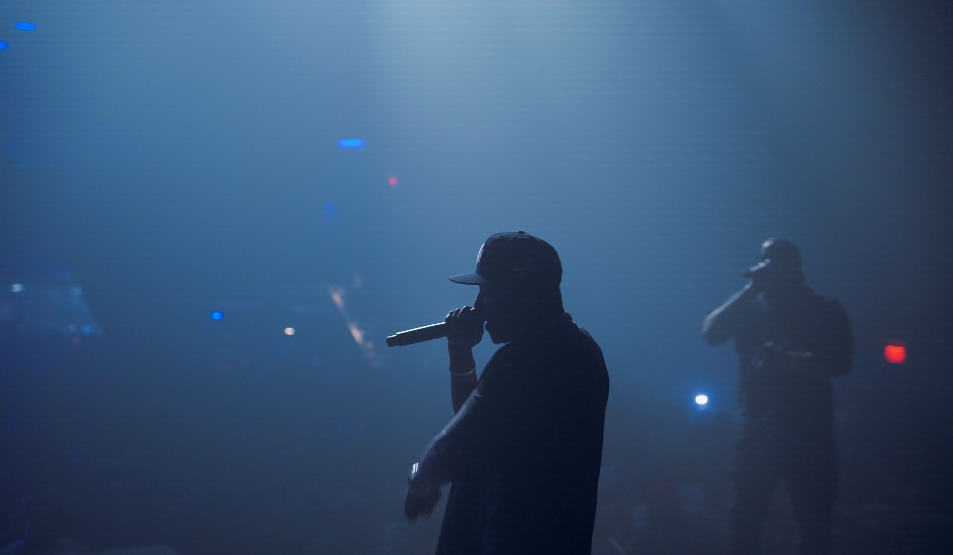 Silhouettes of two performers with microphones on stage in a smoky, blue-lit environment.